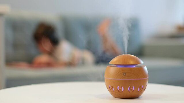 Modern aroma oil diffuser on the white table. Little boy in the background reading a book