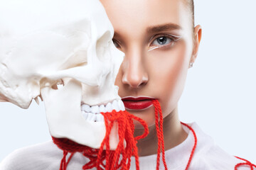 A young beautiful girl with delicate makeup holds a human skull in her hands with a red thread.
