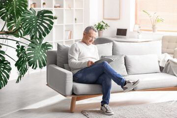 Senior man in eyeglasses reading magazine on sofa at home