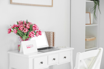 Workplace with modern laptop, beautiful tulips and greeting card with text HAPPY WOMAN'S DAY near light wall in room