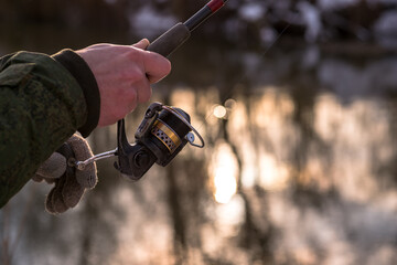 The frozen hands of a male angler holding a spinning reel wind a fishing line on a reel against the background of the river. Selective focus on rod coil. Background