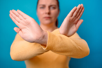 Plakat Angry caucasian 20s woman in yellow sweater crossing hands makes stop gesture, demonstrates rejection isolated over blue background.