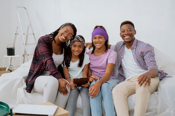 Portrait of a happy smiling loving family sitting on a couch during an apartment renovation. Young parents and two daughters browsing room inspirations on a tablet