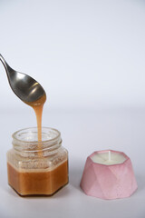 a spoon takes out a berry cream-honey from a jar, next to a candle in a pink plaster candlestick on a white background.