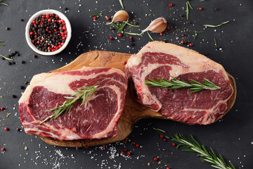 Raw meat, two beef steaks on a cutting board with rosemary and spices on a black background, top...