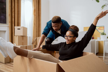 Happy couple first time home buyers having fun while unpacking boxes of laughter on moving day excited wife driving around sitting in cardboard box while husband pushes her around in new apartment - obrazy, fototapety, plakaty
