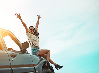 Enjoying life as one whirlwind of an adventure. Portrait of a young woman sitting on a car with her arms outstretched while on a roadtrip.