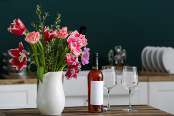 Jug with beautiful flowers, bottle of wine and glasses on table in kitchen