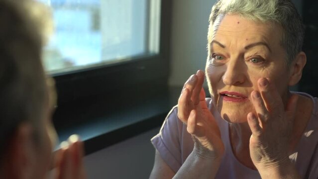 Senior female looking at sagging skin face in mirror, old age appearance. Portrait of senior woman applying anti-aging cream. Portrait of an attractive middle aged woman looking into a mirror