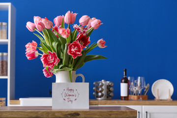 Jug with tulips and greeting card with text HAPPY WOMEN'S DAY on kitchen counter against blue background