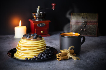 Pancakes with blueberries and chocolate paste on black dish, cup of coffee with steam, coffee...