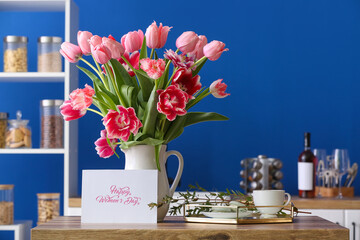 Jug with tulips, cup, macarons and greeting card with text HAPPY WOMEN'S DAY on kitchen counter