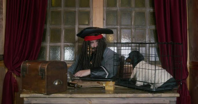 Pirate captain in cocked hat and long coat plays with dressed black dachshund friend sitting IN cage on table in cabin of ship