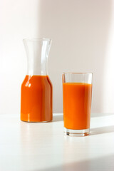 Pitcher with fresh juice on background. Glass of carrot juice on foreground on the white table. Peel and chop the carrots. Place them in a blender, add some water. Blend on high speed