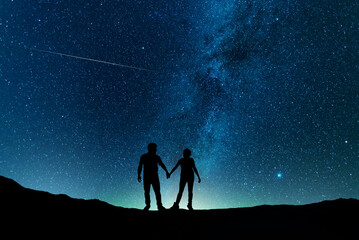 Silhouettes of people in the night. A couple in love is stands on the hill and looks at the bright milky way galaxy. - Powered by Adobe
