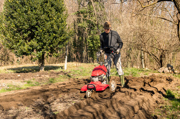 Senior man tilling ground soil with a rototiller in the garden. Spring garden preparation for...