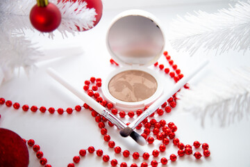A white pillow with powder and bronzer lies on a white background among a white fluffy Christmas tree with red toys, thin makeup brushes and bright red beads lie in front. High quality photo