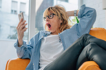 middle aged woman at home looking at mobile phone astonished and surprised