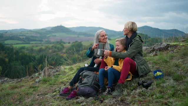 Small girl with mother and grandmother sitting and drinking hot tea on top of mountain.