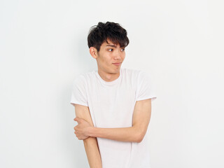 Portrait of handsome Chinese young man with curly black hair in white T-shirt posing against white wall background. Hand on arm and looking away with funny expression, looks frustrated, front view.