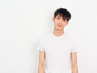Portrait of handsome Chinese young man with curly black hair in white T-shirt posing against white wall background. Arms beside his body and smiling at camera, looks confident, front view.