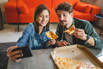 two people young couple man and woman or friends sitting at home at the table taking selfie photos with mobile phone smartphone while eating pizza spending time together having fun bonding in love