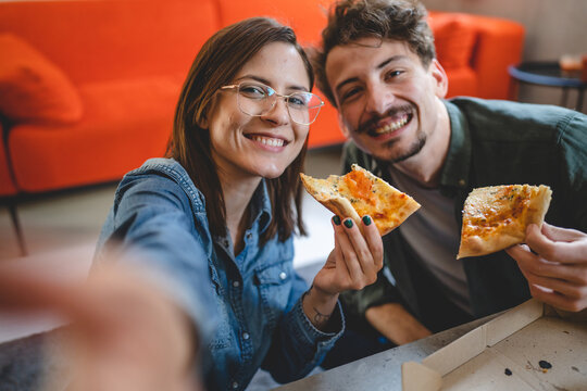 two people young couple man and woman or friends sitting at home at the table taking selfie photos with mobile phone smartphone while eating pizza spending time together having fun bonding in love