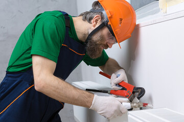 A male plumber installs a radiator in the heating system of an apartment. Guy in overalls and a gas...