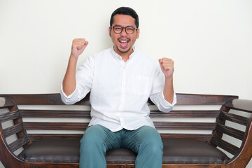 Adult Asian man sitting in a couch showing excited expression with both fist clenched