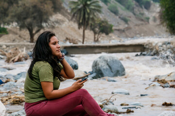 Woman sitting on a river bank holding a cell phone with her hands.