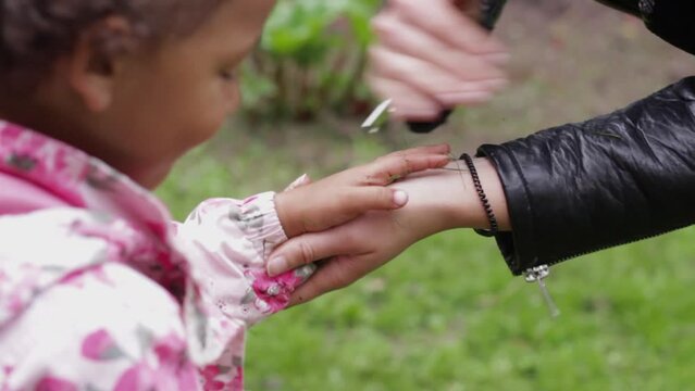 Close up of adult caucasian hands dusting off dirt from little African daughter's hands in pink raincoat after playing outdoor in park, handheld, day