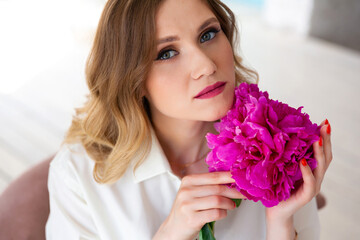 woman in shirt, holding peony bud in her hands, looks at camera. Portrait of beautiful blonde woman. Surprise bouquet of flowers for date, mothers day or valentines day