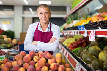 Positive confident seller standing with crossed arms near counter with fresh fruits and vegetables in store..