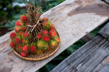 A large bunch of red rambutan in a bamboo tray is placed on a wooden table. Looks delicious to eat rambutan, fresh fruit from the tree in Thailand. Leave space for text