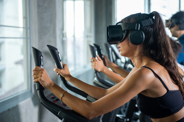 Healthy athletic man and woman wearing VR glasses workout exercise on cycling machine in virtual reality cyberspace at fitness gym. Metaverse digital entertainment technology and sport concept