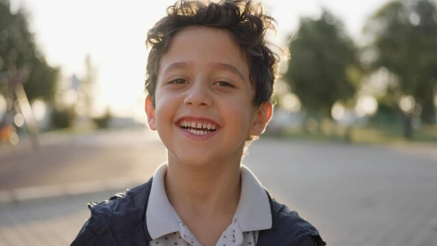 Adorable happy kid baby boy is looking to the camera. Baby looks around and then smiles and laughs. 5 year old fair-haired child with brown eyes smiles at park. Slow motion