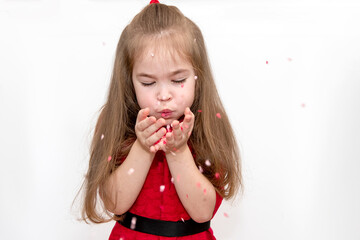 Concept Valentines Day. A girl child with long hair in a red jacket blows on a confetti in her palms.