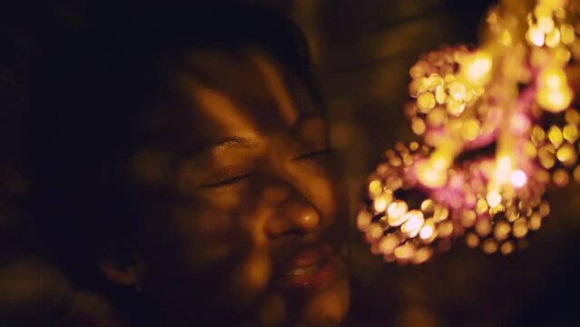 Magic hot light from lanterns falls on the woman eyes. The naked adult sexy caucasian girl lies at night on the bed and holds warm lamps in her hands. Christmas toy in black background. Indoor.