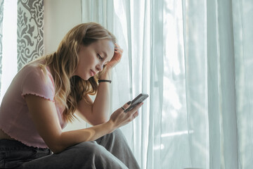 A girl with long hair is sad and reads something on the phone, experiences negative emotions, is upset, sad. Against the background of the window of the house, during the day