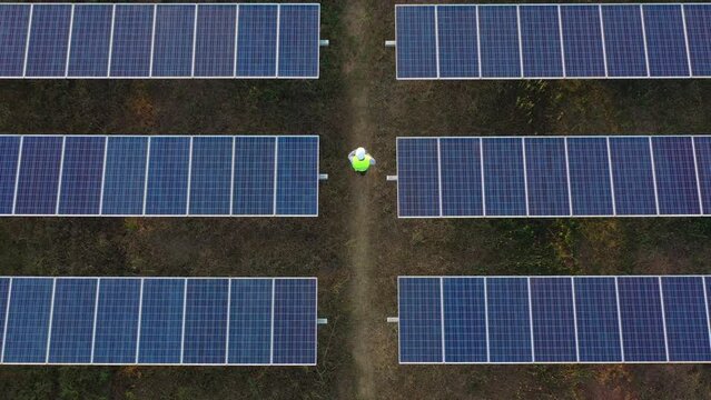 Aerial drone view, Flight over solar panel farm and young engineer wearing white helmet talking on smartphone and walking between solar panels row