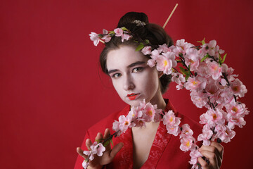 Image beautiful young Geisha woman in traditional Japanese kimono isolated on a red background holding a branch of sakura in her hands. Caucasian woman in Chinese woman make-up