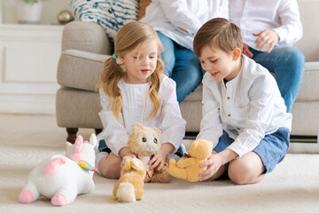 Cute little brother and sister sitting on floor playing with soft toys, parents relaxing on a comfortable sofa in the living room young caucasian family relaxing together enjoying the weekend at home