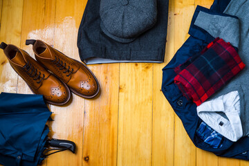 Clothing Concepts. View Flat Lay of Tan Brogues Boots With Umbrella, Jumper, Checked Winter Scarf, Cap, Herringbone Trousers, White Shirt and Raincoat on Wooden Surface Background.