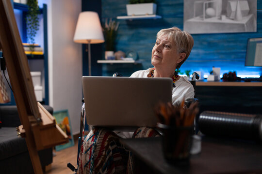 Older person sitting in wheelchair holding laptop looking at pencil sketch drawing on easel. Elderly woman artist living with disability using portable computer looking for inspiration.