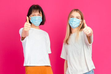 Couple of Two Positive Young Girlfriends Demonstrating Application of Facial Protection Surgical Mask With Thumbs Up Gesture Against Pink Background