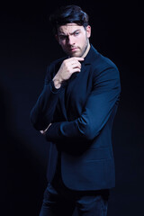 Thinking Caucasian Brunet Man In Black Suit Posing With Folded Hands Looking To Side Over Dark Background in Studio.