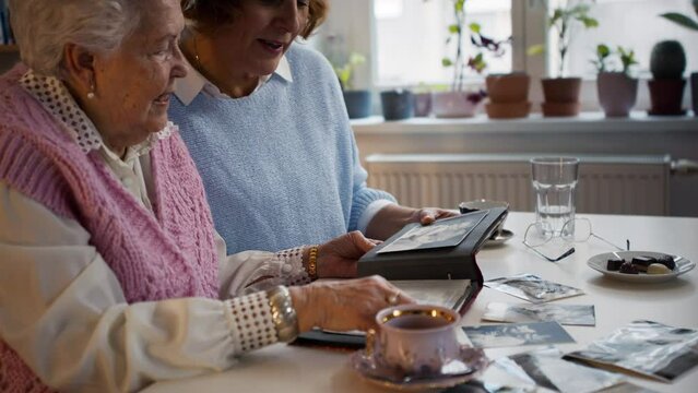 Senior mother with her adult daughter looking at old family pictures at home together