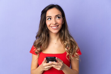 Fototapeta premium Young caucasian woman isolated on purple background using mobile phone and looking up