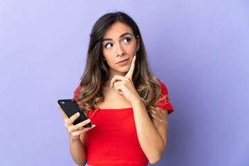 Naklejka premium Young caucasian woman isolated on purple background using mobile phone and thinking