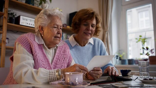 Senior mother with her adult daughter looking at old family pictures at home together